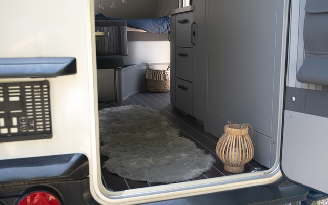 Offene Hecktür eines Wohnmobils, Blick ins Innere mit grauem Schrank, oberem Bett mit blauem Bezug, rundem hellgrauen Teppich und geflochtener Laterne auf dem Boden