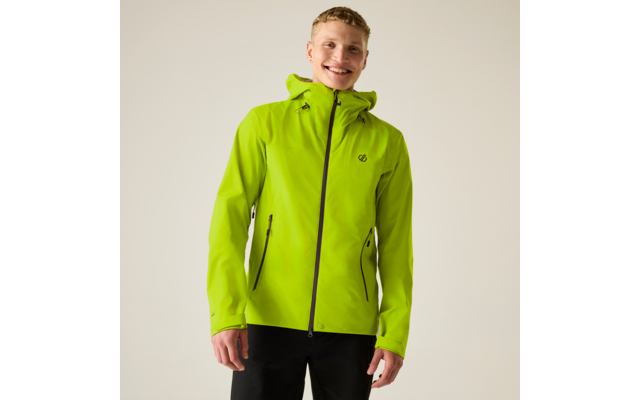 Person mit kurzem blondem Haar trägt hellgrüne Jacke mit Kapuze und schwarze Hosen, steht vor hellem Hintergrund und lächelt