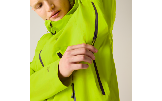 Teilfoto einer Person, die den linken Arm hebt und mit der Hand einen senkrechten schwarzen Reißverschluss an einer neongrünen Jacke berührt; Teil des Gesichts, Hals und Hand sichtbar, neutraler Hintergrund