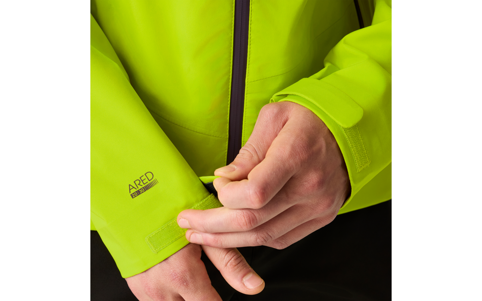 Hände, die den Klettverschluss an der Ärmelmanschette einer leuchtend grünen Jacke mit Reißverschluss einstellen; kleines ARED-Logo auf dem Ärmel