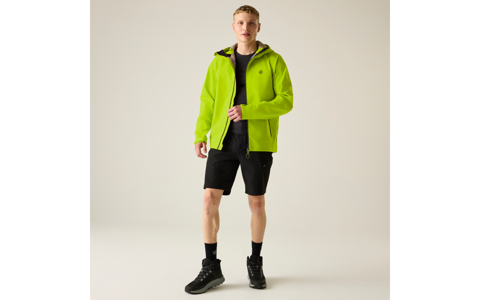 Stehende Person mit kurzem blondem Haar, trägt neongrüne Kapuzenjacke, schwarzes T-Shirt, schwarze Shorts, schwarze Socken und schwarze Sportschuhe vor hellem einfarbigem Hintergrund