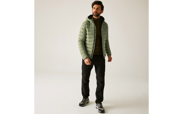 Ein Mann trägt eine grüne Steppjacke, schwarzen Hose und schwarze Schuhe, stehend vor hellem Hintergrund