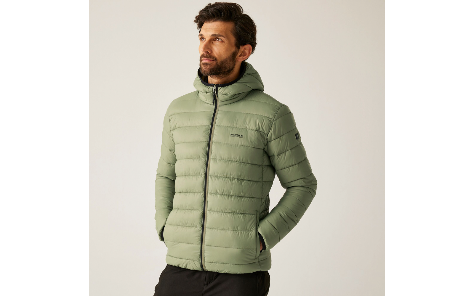 Mann mit kurzem braunem Haar und Bart trägt eine grüne Steppjacke und schwarze Hose vor hellem Hintergrund