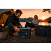 Homme à genoux près d'une centrale électrique portable en train de la manipuler, femme assise sur une chaise de camping tenant une tasse, en arrière-plan tente, lanterne, lac, montagnes et coucher de soleil