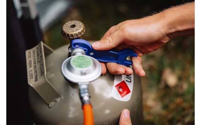 Hände montieren einen Druckregler mit blauem Spannwerkzeug an eine kleine graue Gasflasche, oranger Gasschlauch sichtbar