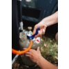 Zwei Hände verbinden einen orangen Schlauch mit einer grauen Gasflasche und blauem Regler; offene Autotür und Gras im Hintergrund