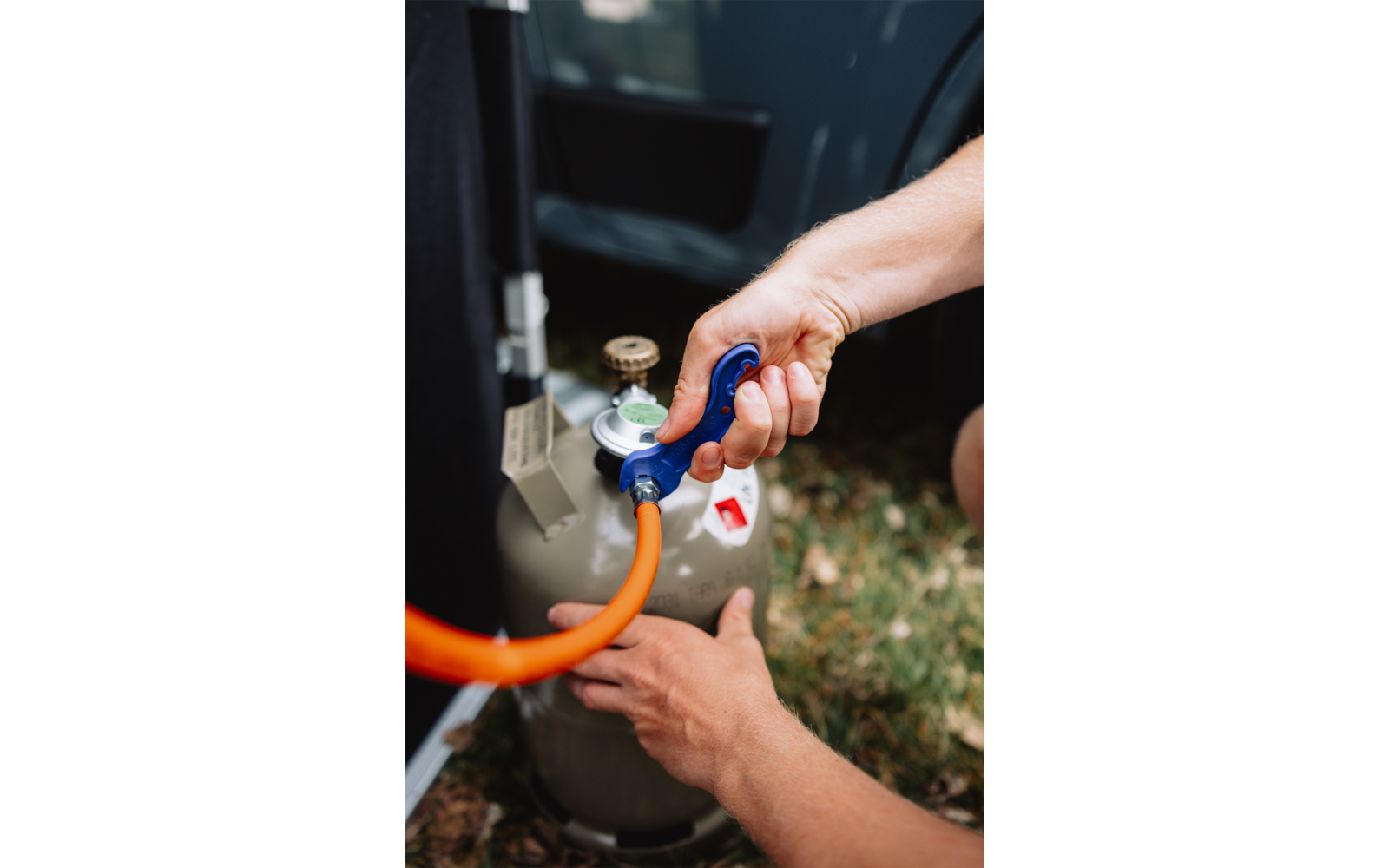 Zwei Hände verbinden einen orangen Schlauch mit einer grauen Gasflasche und blauem Regler; offene Autotür und Gras im Hintergrund
