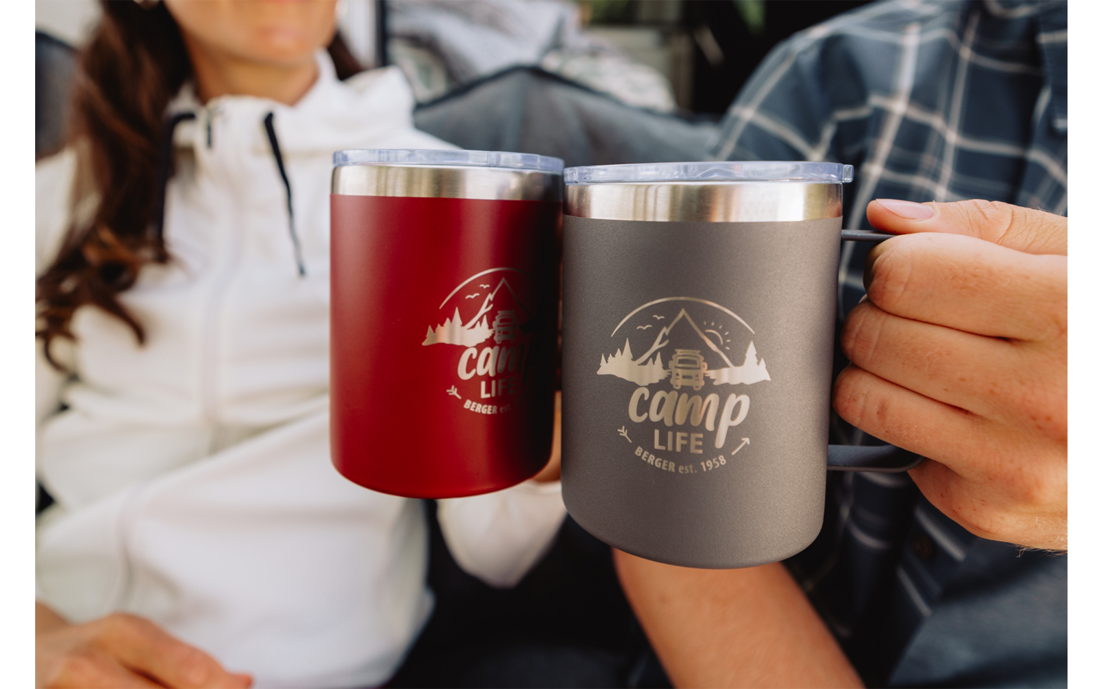 Twee personen zitten naast elkaar, elk houdt een geïsoleerde beker met RVS-rand en het logo 'Camp Life' vast; één beker rood, één grijs. Gezichten niet volledig zichtbaar