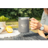 Main tenant une tasse grise à bord métallique avec inscription 'camp life' sur une table en bois ; moitié de citron, bol blanc de baies, petite plante en pot, bras avec t-shirt rayé et montre, arrière-plan vert flou
