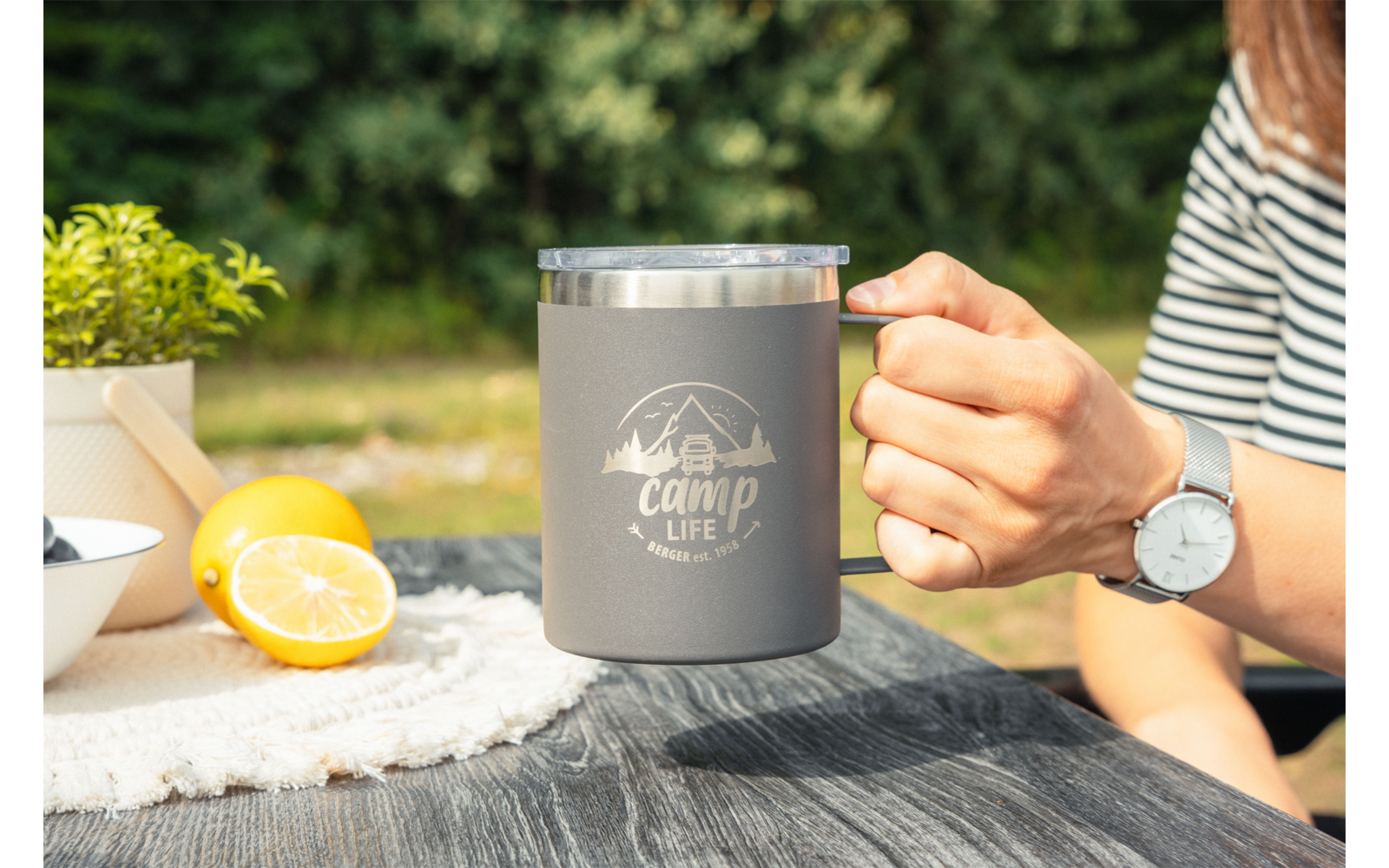 Main tenant une tasse grise à bord métallique avec inscription 'camp life' sur une table en bois ; moitié de citron, bol blanc de baies, petite plante en pot, bras avec t-shirt rayé et montre, arrière-plan vert flou