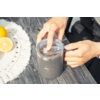 Deux mains ouvrent le couvercle d'une tasse isotherme grise posée sur une table en bois; à gauche, deux moitiés de citron sur un dessous de tasse blanc tissé