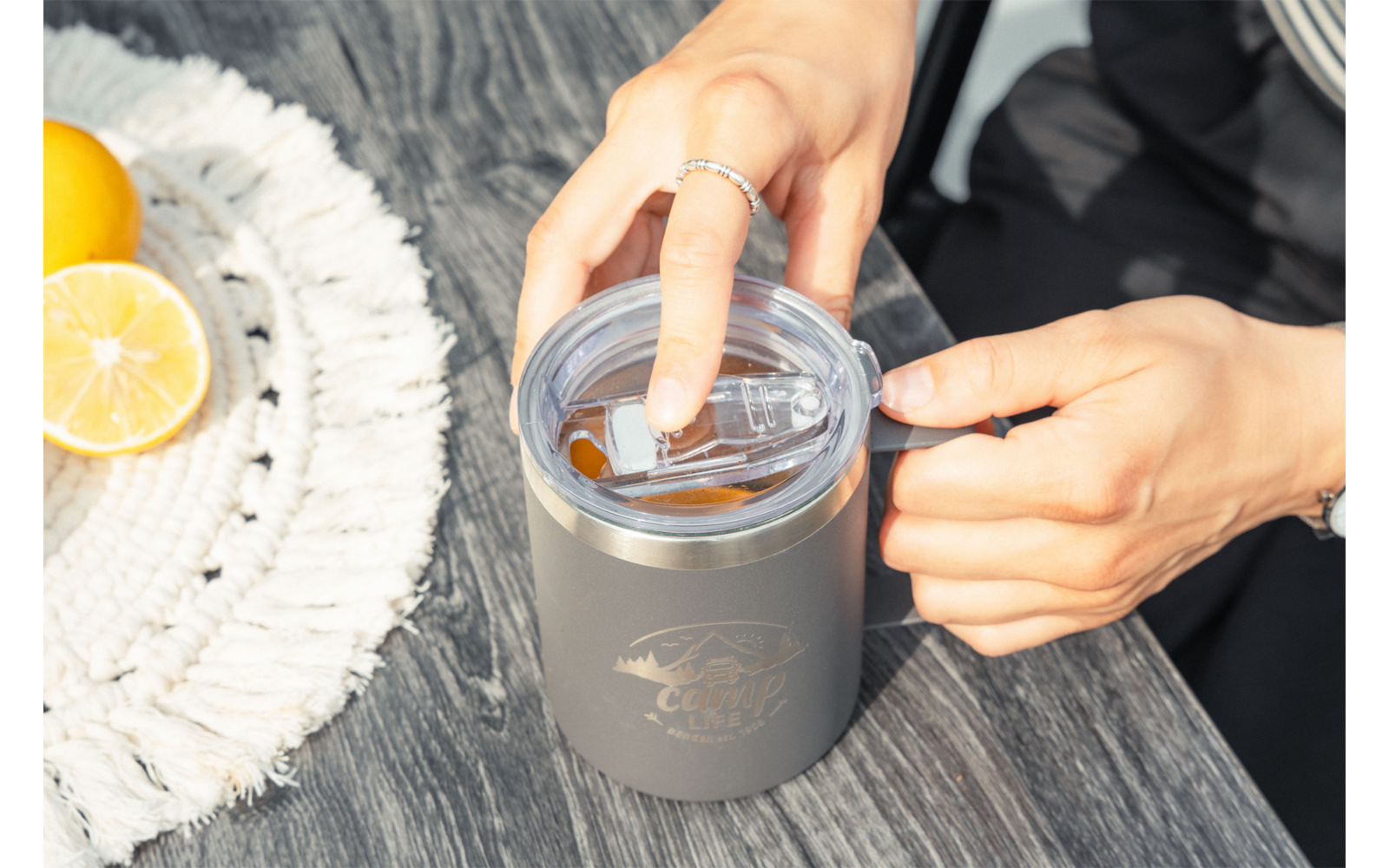 Deux mains ouvrent le couvercle d'une tasse isotherme grise posée sur une table en bois; à gauche, deux moitiés de citron sur un dessous de tasse blanc tissé