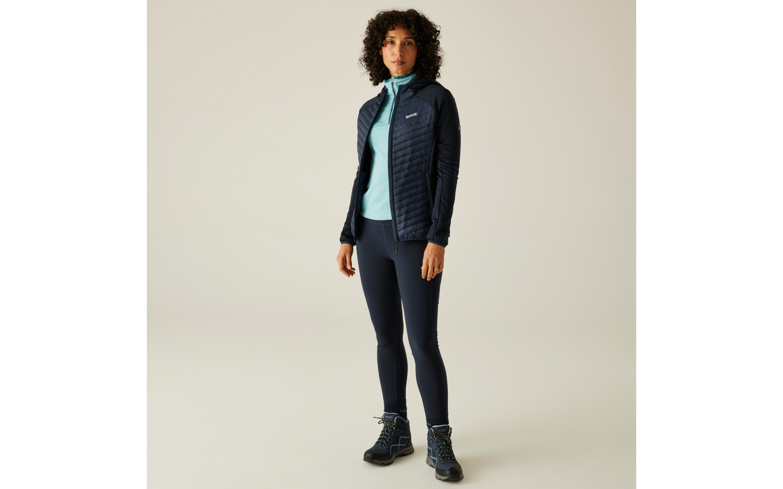 Person mit lockigen dunklen Haaren trägt dunkle gesteppte Jacke, hellblaues Oberteil, dunkle Leggings und blaue Wanderschuhe, steht vor hellem Hintergrund