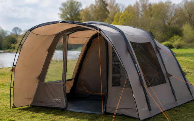 Grande tente tunnel gris-beige avec entrée ouverte laissant voir l'intérieur, haubans orange, installée sur l'herbe près d'un lac et d'arbres sous un ciel nuageux