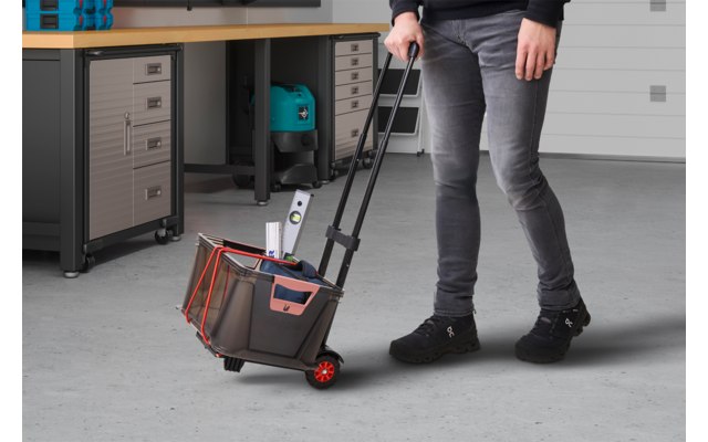 Personne tirant une caisse à outils roulante avec poignée télescopique et roulettes rouges dans un atelier; établi avec tiroirs et un appareil en arrière‑plan