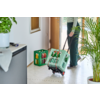 Bas du corps d'une personne dans l'encadrement d'une porte ouverte, tirant une caisse verte à roulettes remplie de bouteilles en verre ; une autre caisse verte sur le sol carrelé du couloir ; plantes en pot et meuble à chaussures visibles