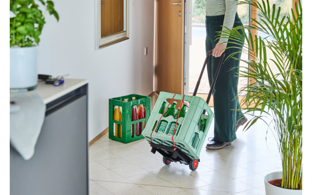 Bas du corps d'une personne dans l'encadrement d'une porte ouverte, tirant une caisse verte à roulettes remplie de bouteilles en verre ; une autre caisse verte sur le sol carrelé du couloir ; plantes en pot et meuble à chaussures visibles