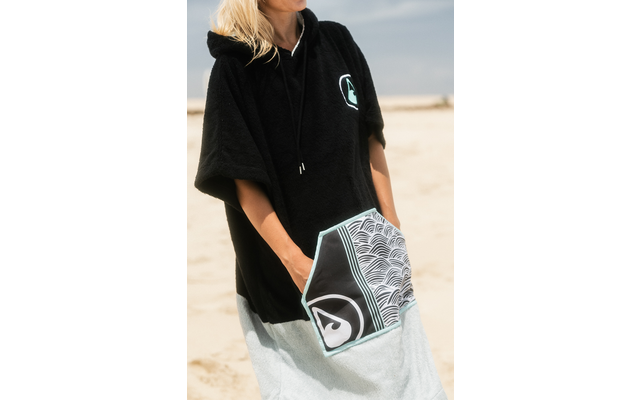Torso de una persona en una playa de arena que lleva un poncho negro con capucha y un bolsillo delantero con patrón de ondas; manos en el bolsillo, cabello rubio visible