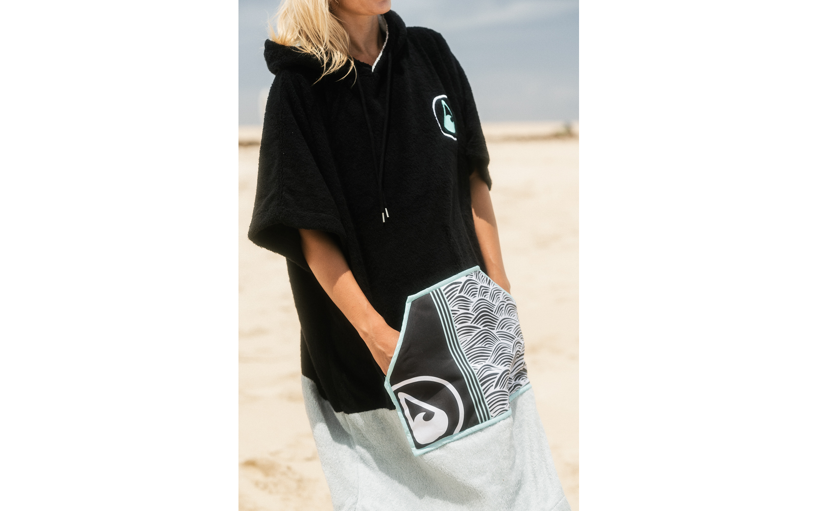 Torso de una persona en una playa de arena que lleva un poncho negro con capucha y un bolsillo delantero con patrón de ondas; manos en el bolsillo, cabello rubio visible