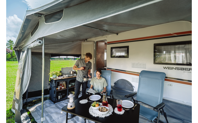 Zwei Personen, eine sitzt, eine steht, an einem Campingtisch mit Tellern, einer Schüssel und Gläsern vor einem Wohnwagen unter einer Markise