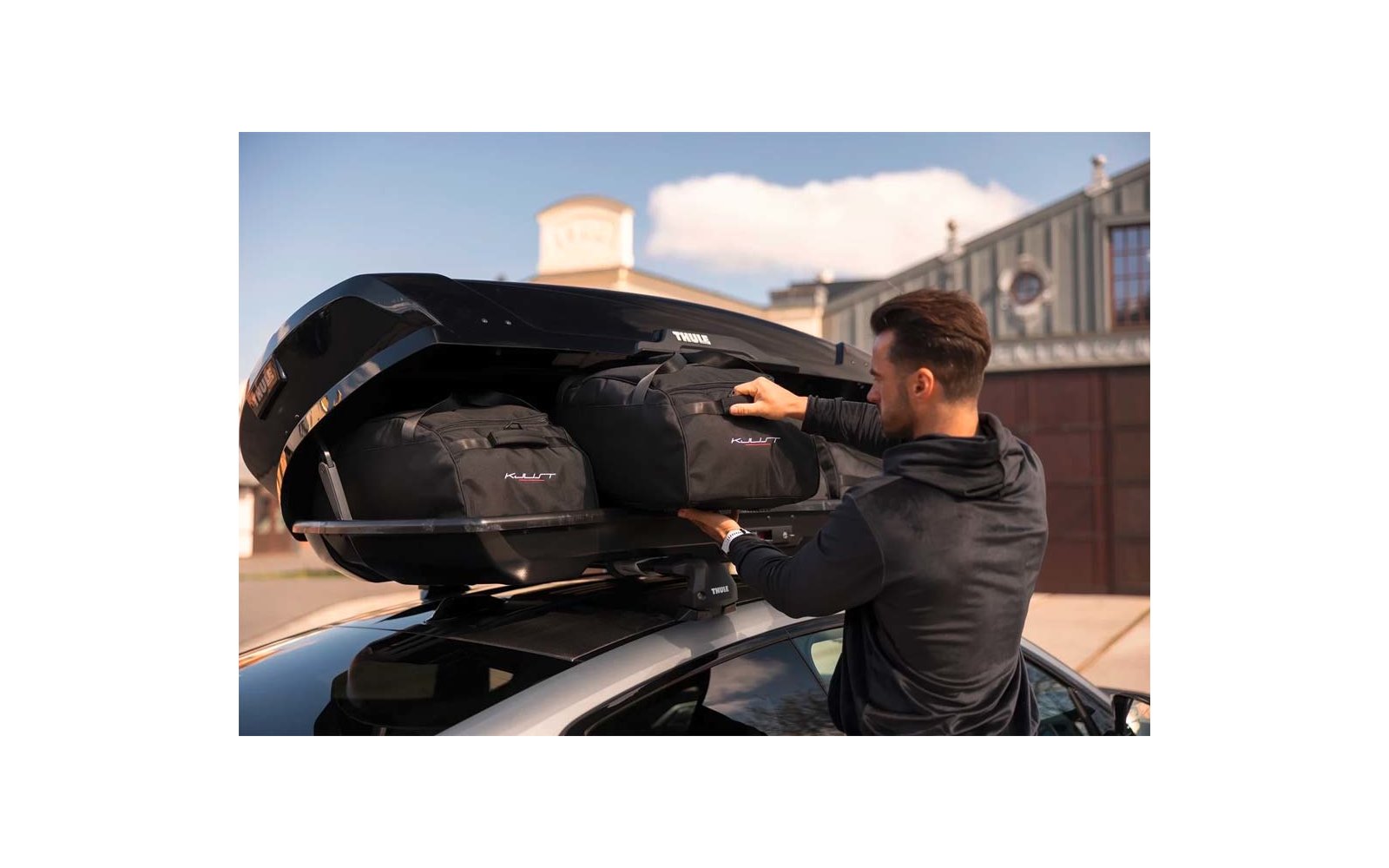 Mann legt schwarze Reisetaschen in eine geöffnete Dachbox auf einem Auto, Gebäude im Hintergrund