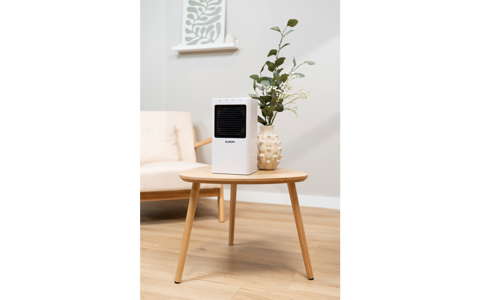 Weißes rechteckiges Gerät mit schwarzer Front auf rundem Holztisch, daneben eine beigefarbene Vase mit grünen Zweigen, im Hintergrund ein heller Sessel mit Kissen, Wandablage mit dekorativem Paneel, heller Holzfußboden