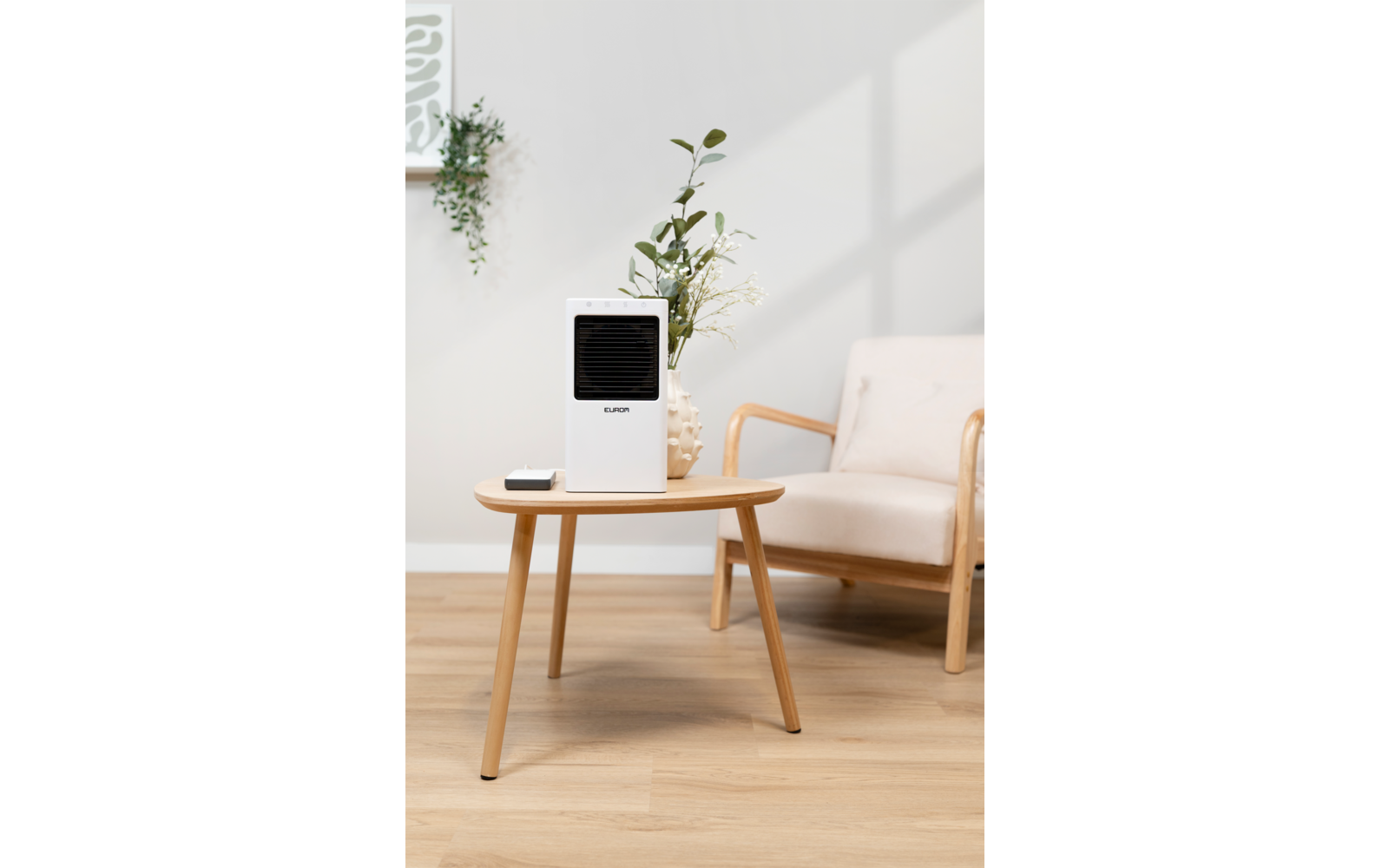 Weißes rechteckiges Gerät mit schwarzer Front auf rundem Holztisch; Smartphone auf dem Tisch; Vase mit Zweigen dahinter; beige Sitzgelegenheit rechts; heller Raum mit Holzboden