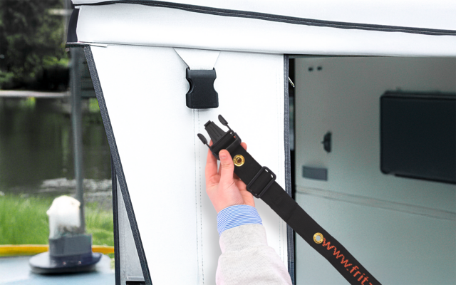 Hand mit hellem Ärmel befestigt schwarzen Gurt mit Steckschloss an weißem Stoffpanel; im Hintergrund Wasser, Gras und ein Poller