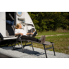 Persoon zit in een ligstoel met uitgestrekte benen op een voetensteun naast een witte caravan met geopende deur, op een patroonmat op gras, bomen op de achtergrond