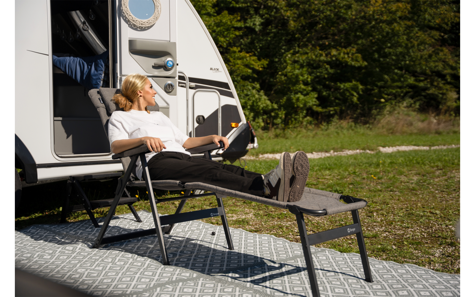 Persoon zit in een ligstoel met uitgestrekte benen op een voetensteun naast een witte caravan met geopende deur, op een patroonmat op gras, bomen op de achtergrond