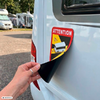 Hand met twee ringen die een rode sticker met "ATTENTION" en een bestelwagen-icoon van de achterkant van een witte camper lostrekt; op de achtergrond andere campers en bomen