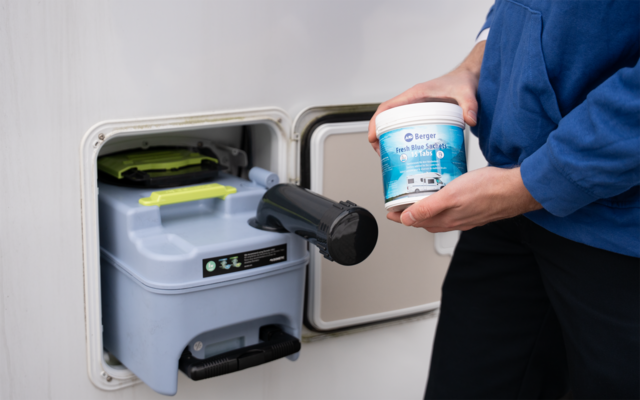 Offenes Außenfach eines Wohnwagens mit herausgezogener grauer Kunststoffkassette und schwarzem Ausgussrohr; Person in blauem Oberteil hält weiße Plastikdose mit blauem Etikett; grüner Deckel auf der Kassette sichtbar