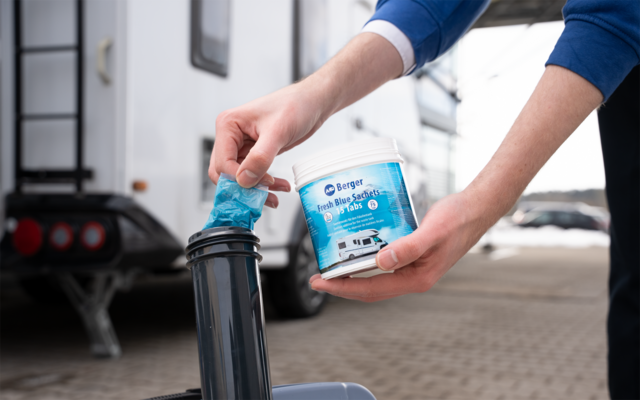 Hände einer Person, links ein blaues Tuch in einer schwarzen runden Öffnung, rechts eine weiße Dose mit blauem Etikett; im Hintergrund ein geparkter Wohnwagen