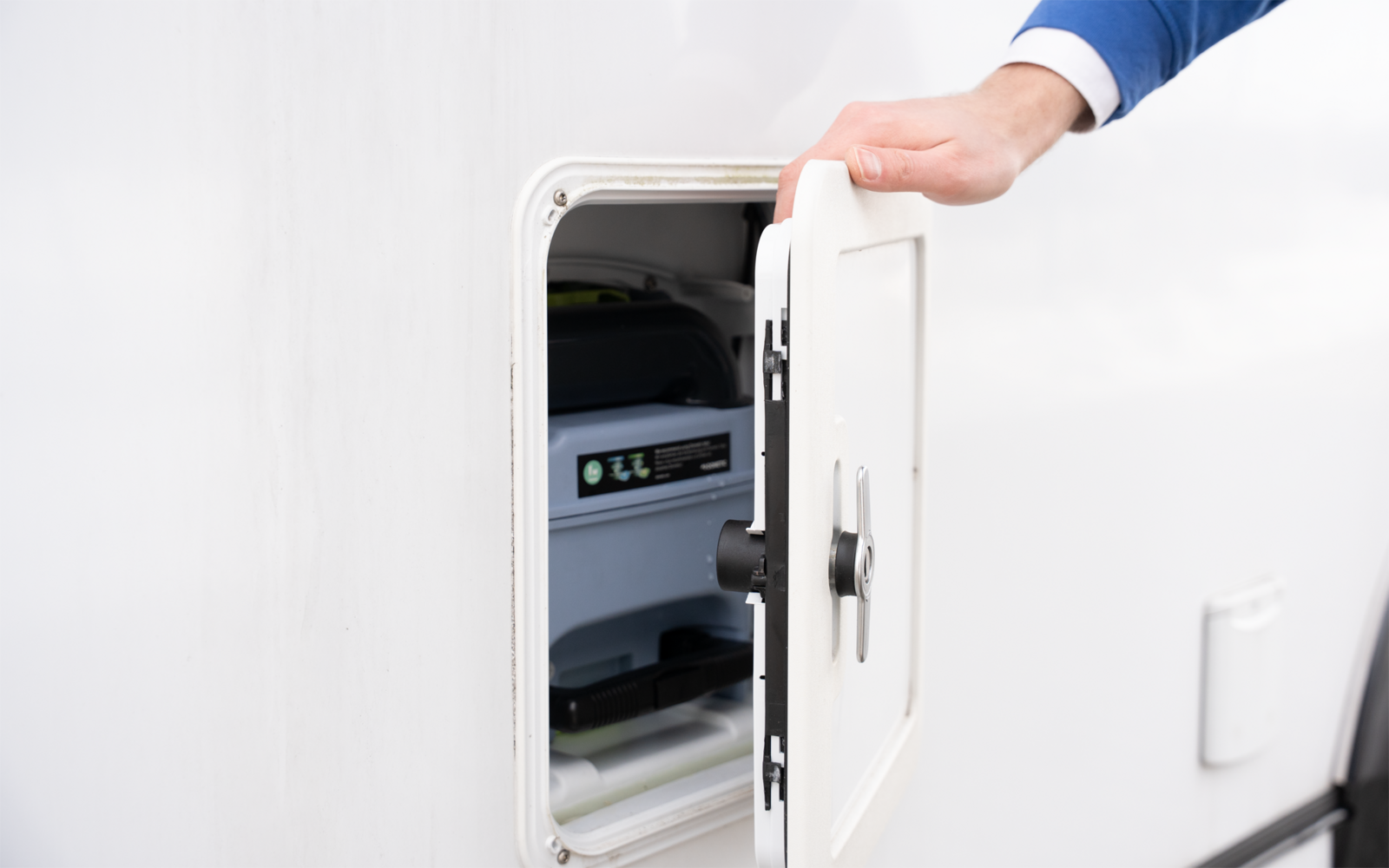 Hand in blauem Ärmel öffnet weiße Außenklappe eines Fahrzeugs; innen ein Gerät mit Display und Kabeln