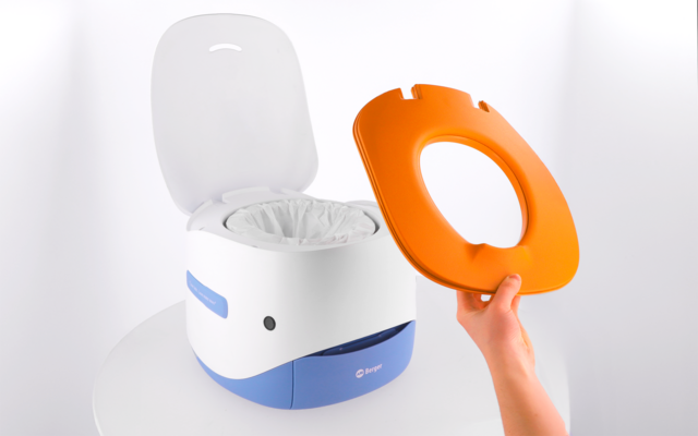 Weiß-blaues Kunststoffgerät mit geöffnetem Deckel, weiße Einwegfolie innen sichtbar; eine Hand hält einen orangefarbenen Toilettensitz darüber; Gerät steht auf weißer Oberfläche vor weißem Hintergrund