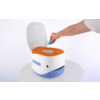 Kleine tragbare Toilette weiß mit orangefarbenem Sitz und blauem Sockel auf einem runden Tisch; eine Hand legt einen blauen Beutel in die Toilette