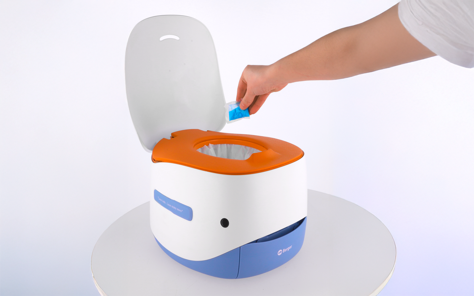 Kleine tragbare Toilette weiß mit orangefarbenem Sitz und blauem Sockel auf einem runden Tisch; eine Hand legt einen blauen Beutel in die Toilette