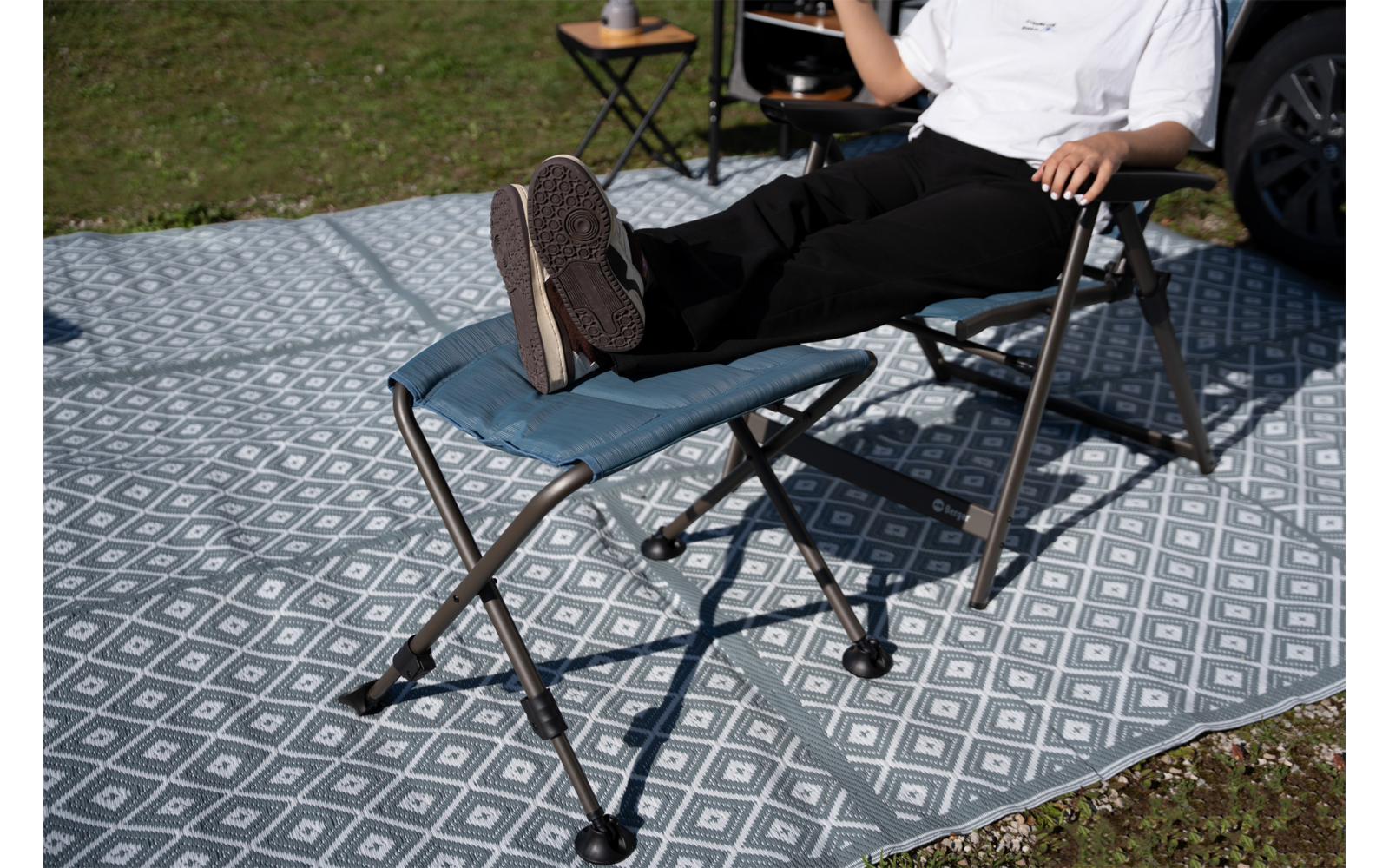 Teilweise sichtbare Person sitzt in Klappstuhl, Beine auf Fußhocker; weißes T‑Shirt, schwarze Hose, Sneaker; gemusterter Outdoor-Teppich, kleiner Beistelltisch mit Tasse, Fahrzeugrad am Bildrand, Gras