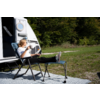 Person mit Sonnenbrille sitzt auf Campingstuhl neben weißem Wohnmobil, Beine auf einem Hocker, hält eine Tasse; Wiese und Bäume im Hintergrund