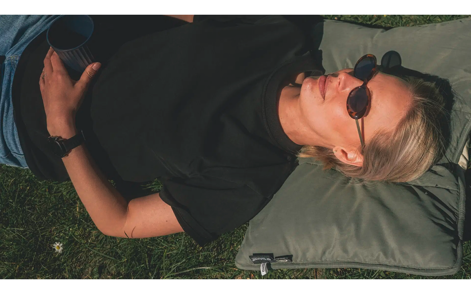 Vrouw met zonnebril liggend op een kussen in het gras, houdt een beker vast