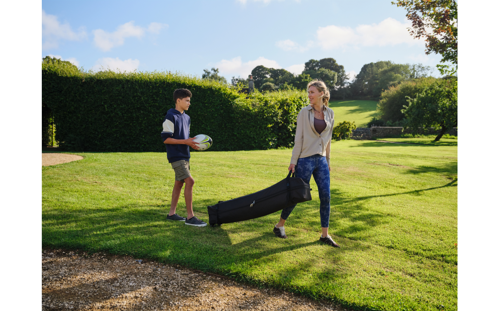 Frau trägt eine lange schwarze Tasche in der Hand, Junge hält weißen und grünen Ball; beide gehen auf einer Grasfläche mit Hecke, Bäumen und blauem Himmel