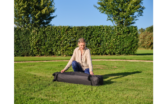 Person kniet auf dem Rasen und hantiert mit einem langen schwarzen Transportbeutel; im Hintergrund Hecke, Bäume und blauer Himmel