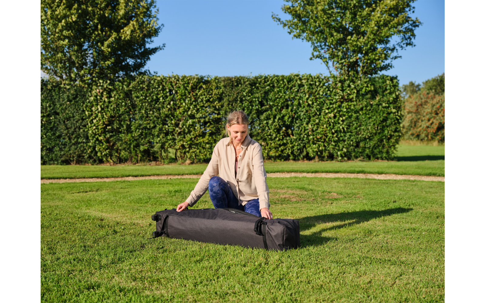Person kniet auf dem Rasen und hantiert mit einem langen schwarzen Transportbeutel; im Hintergrund Hecke, Bäume und blauer Himmel