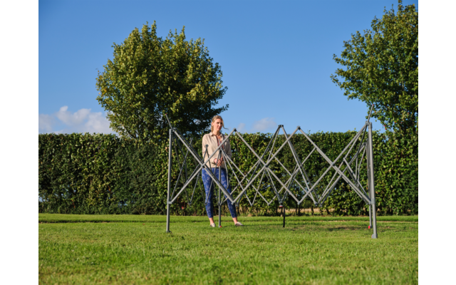 Person steht hinter zusammengeklapptem Metallwäscheständer auf Rasen; im Hintergrund Hecke, zwei Bäume und blauer Himmel