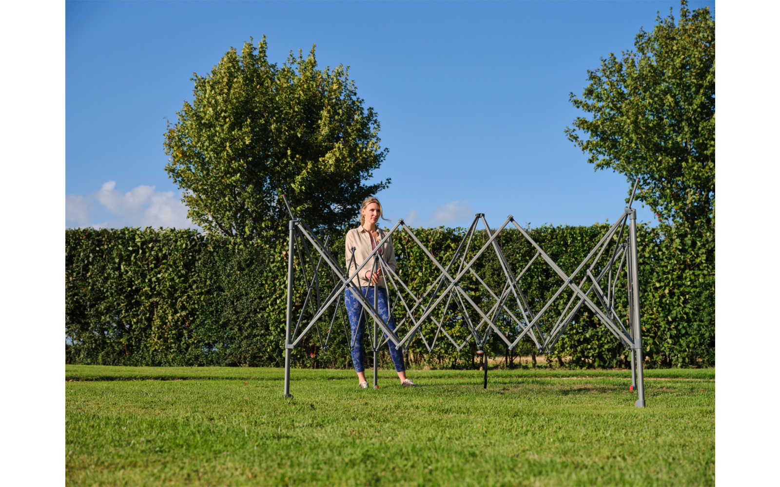 Person steht hinter zusammengeklapptem Metallwäscheständer auf Rasen; im Hintergrund Hecke, zwei Bäume und blauer Himmel