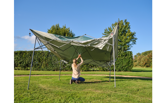Person kniet auf Gras unter einem teilweise zusammengefallenen grünen Pavillon und hält die Bänder des Gestänges; im Hintergrund Hecke, Bäume und blauer Himmel