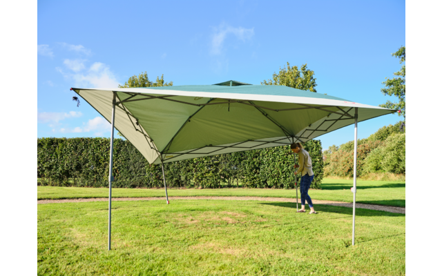 Grünes Faltzelt mit vier Metallstützen auf Rasen, Person stellt eine Stange ein, Hecke und Bäume im Hintergrund, blauer Himmel