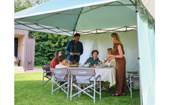 Sieben Personen sitzen und stehen an einem gedeckten Tisch unter einem hellgrünen Pavillon auf einer Rasenfläche; Teller, Getränke und Schalen auf dem Tisch; Haus im Hintergrund