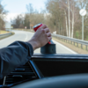 Hand hält rote Dose im Getränkehalter eines Autos, Blick durch die Windschutzscheibe auf eine Straße mit Leitplanke und Bäumen