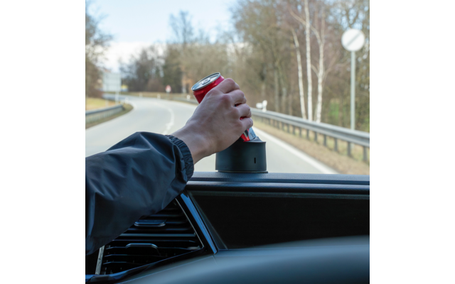 Hand hält rote Dose im Getränkehalter eines Autos, Blick durch die Windschutzscheibe auf eine Straße mit Leitplanke und Bäumen